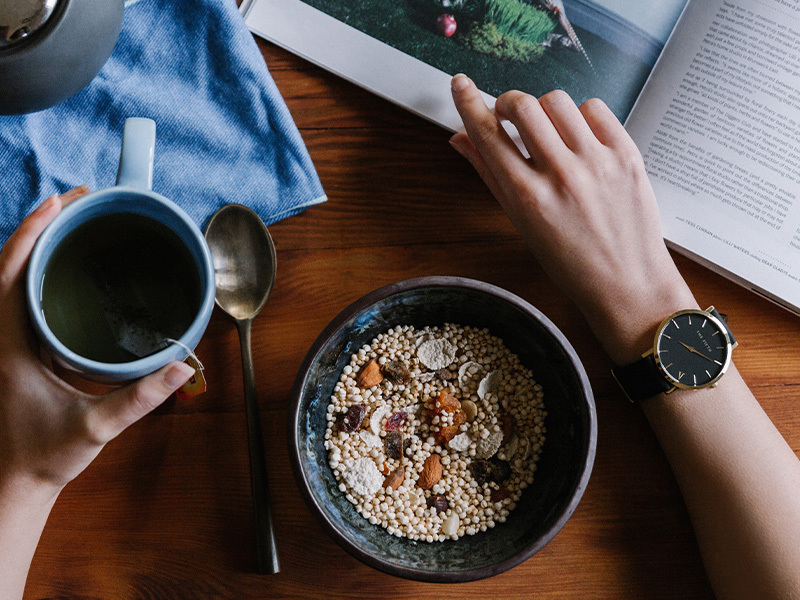 A hand holding a mug of tea, whilst eating breakfast and flicking through a magazine